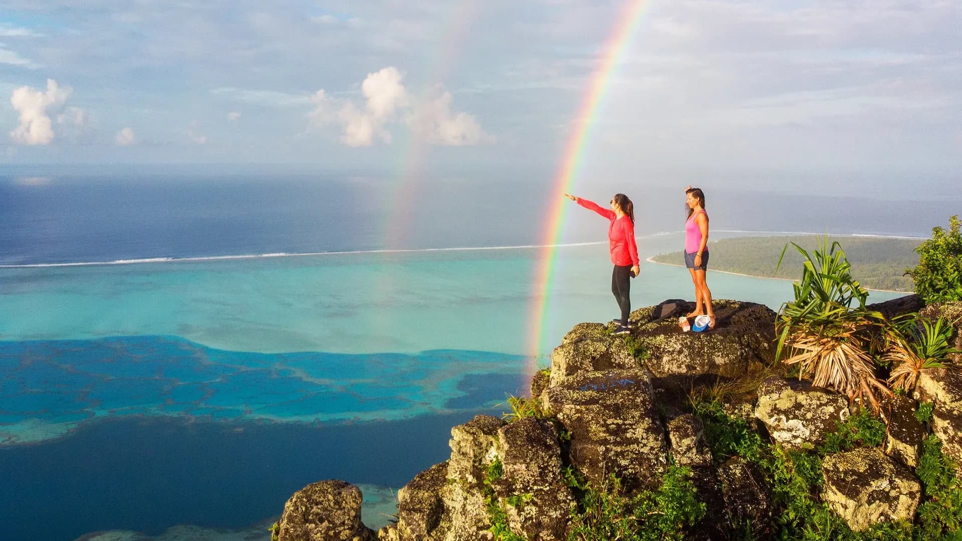 Two Sailing Virgins sailors on a clifftop above the lagoon under a rainbow